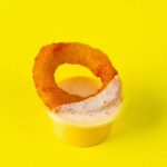 A crispy onion ring partially dipped in creamy sauce against a bright yellow background.